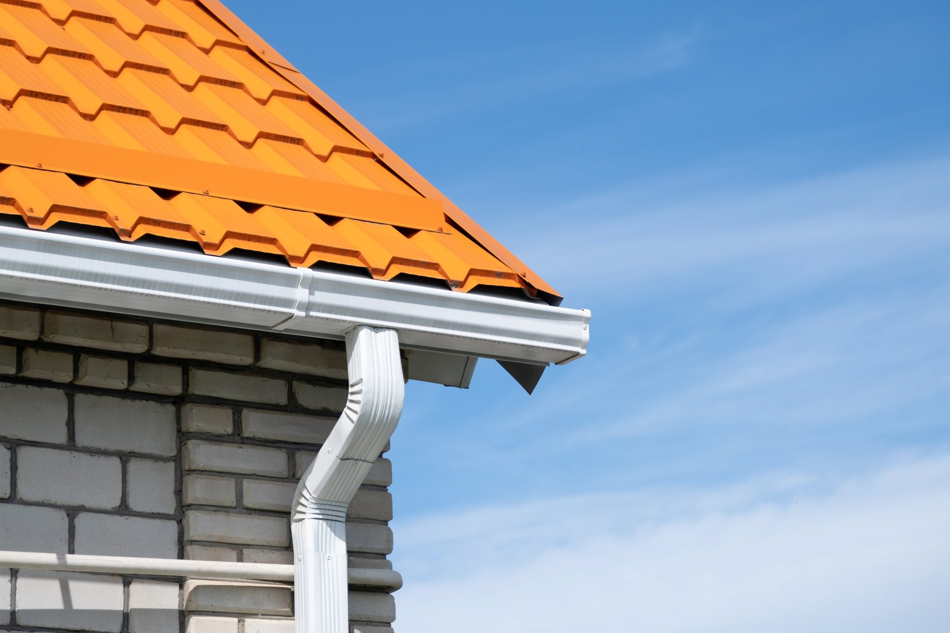 Rain gutter pipeline system on brick house with orange metal tile roof against blue sky